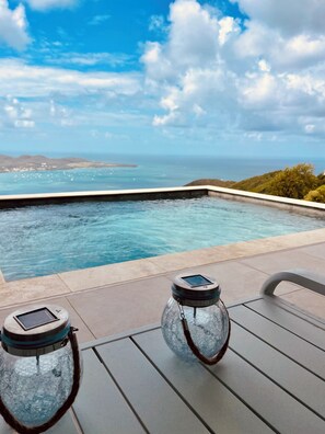 Uma área à beira da piscina com vista clara para o oceano e colinas ao fundo.