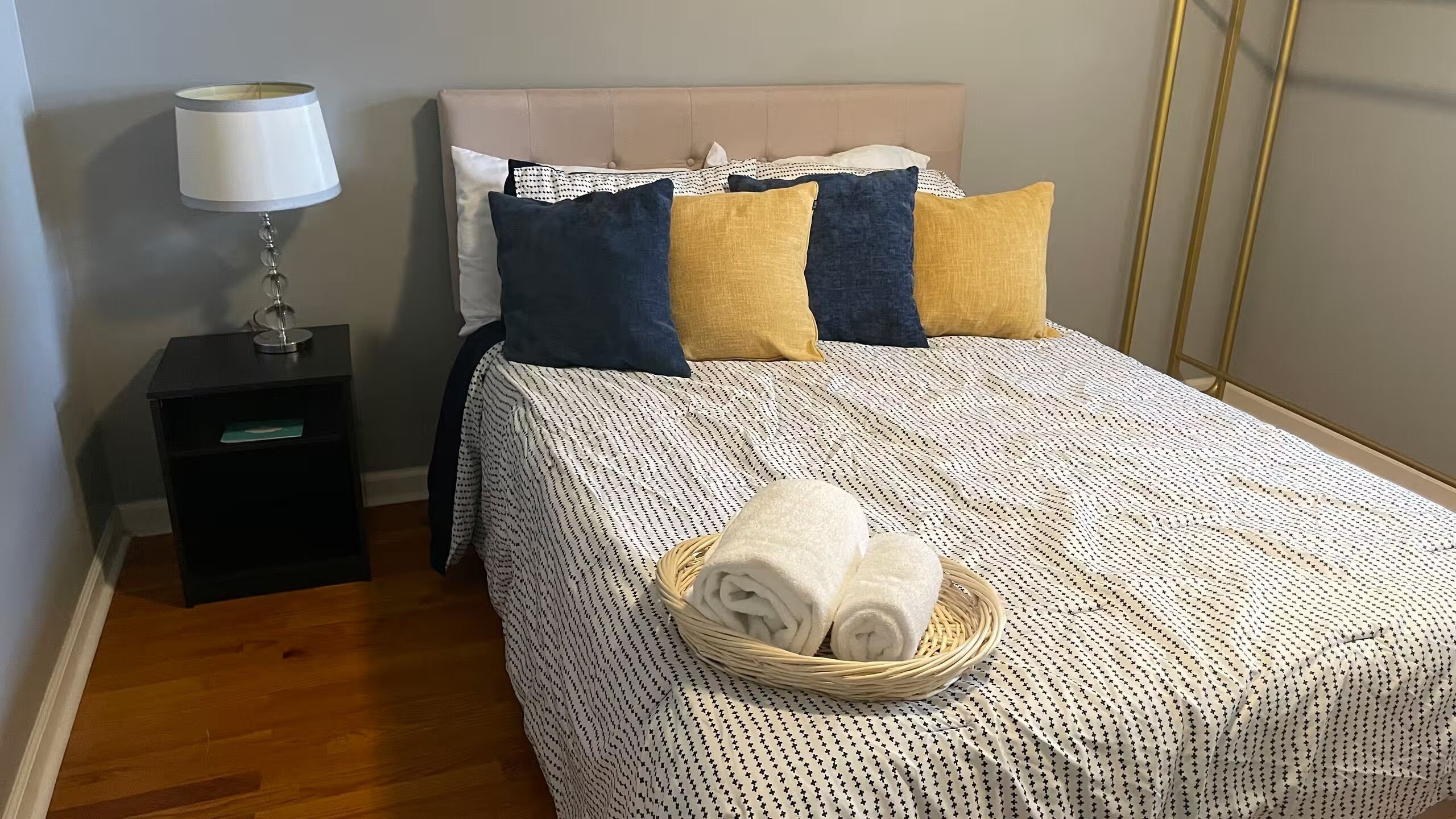 A neatly made bed with a patterned bedspread, a bedside lamp, and a basket of folded towels.