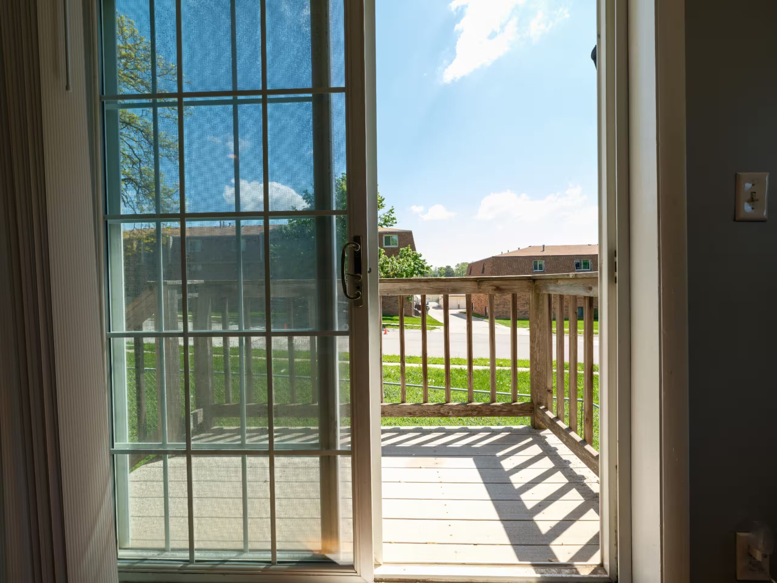 A view through a glass door with a metal grid, leading to an outdoor area with a wooden deck and a brick building in the background.
