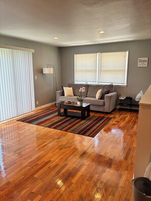 A living room with a wooden floor, a sofa, a coffee table, and a window with blinds.
