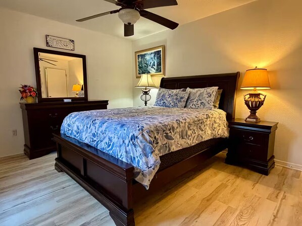 A bedroom with a bed, a nightstand, a lamp, a ceiling fan, a mirror, and a framed picture on the wall.