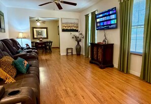 A living room with a leather couch, wooden flooring, a television mounted on the wall, and a dining area in the background.