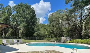 Piscina con sillas de descanso y zona vallada, rodeada de árboles.