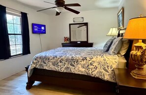 Un dormitorio con cama, ventilador de techo, televisor, ventana con cortinas, mesita de noche con lámpara y un cuadro en la pared.
