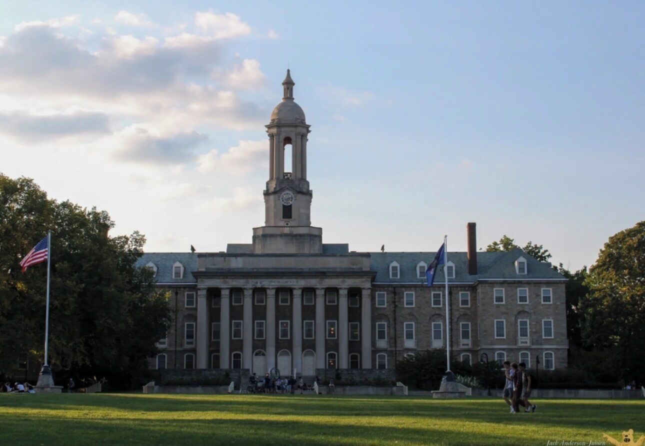 A large building with a prominent clock tower, a flag, and a grassy area in front.