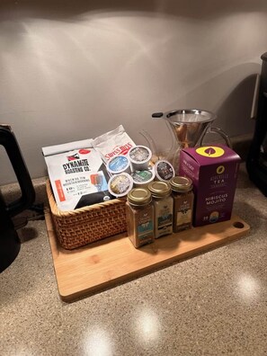 A kitchen counter with a basket containing a bag of Dynamite Roasting Co. Mountain Morning coffee, a box of Ashville Tea Hibiscus Mojito, and several jars of spices. - Dog-Friendly Mountain Home w/ Hot Tub & Rec Room (Asheville)