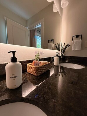 A bathroom with a dark marble countertop, a white sink, a mirror, a bottle of hand soap, a small basket with toiletries, and a potted plant. - Dog-Friendly Mountain Home w/ Hot Tub & Rec Room (Asheville)