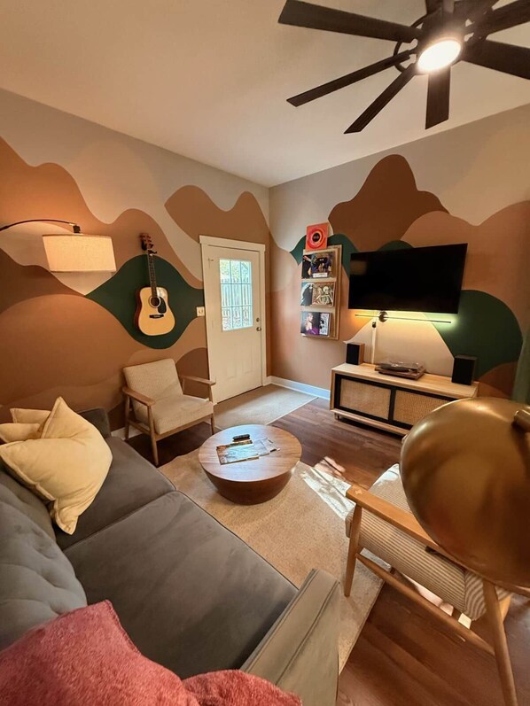 A living room with a sofa, a coffee table, a guitar, a bookshelf, and a television. - Dog-Friendly Mountain Home w/ Hot Tub & Rec Room (Asheville)