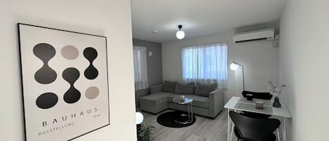 A modern living room with a grey sofa, a small round table, a desk with a chair, and a framed artwork on the wall.