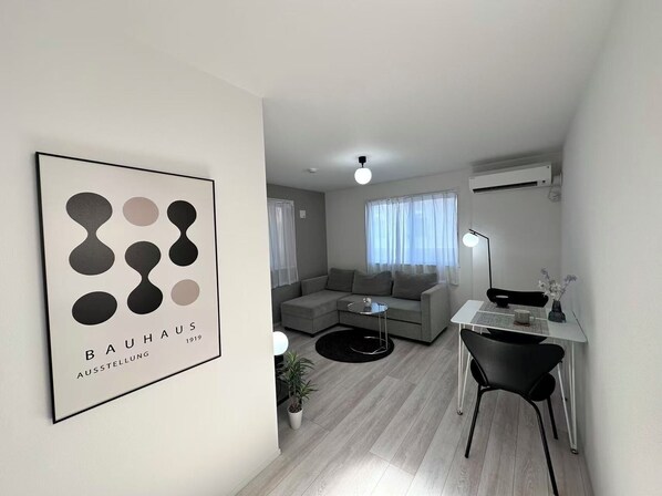 A modern living room with a grey sofa, a small round table, a desk with a chair, and a framed artwork on the wall.