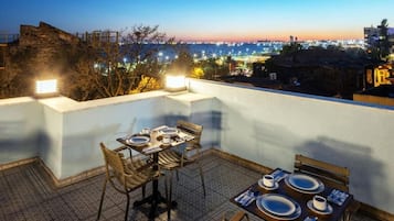 Terraza en la azotea con mesas dispuestas para comer, con vistas a un paisaje urbano al atardecer.