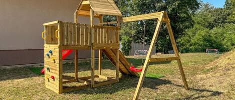 A wooden play structure with a slide and climbing area, set on a grassy lawn.
