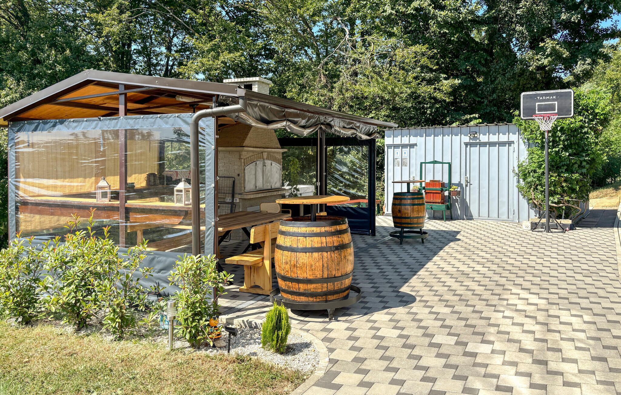 A covered outdoor seating area with a table and chairs, a wooden barrel, and a basketball hoop.
