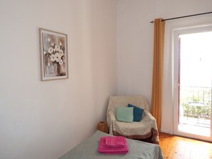 Une pièce comprenant un lit, un fauteuil avec des coussins, une fenêtre avec des rideaux et un tableau encadré accroché au mur.