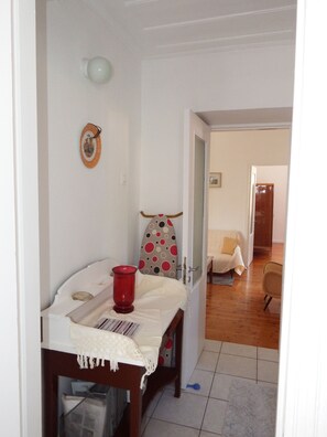 A room with a wooden table, a red candle holder, a patterned cushion, and a doorway leading to another room with a bed and a chair. - Nandia's Molyvos House (Mithymna)