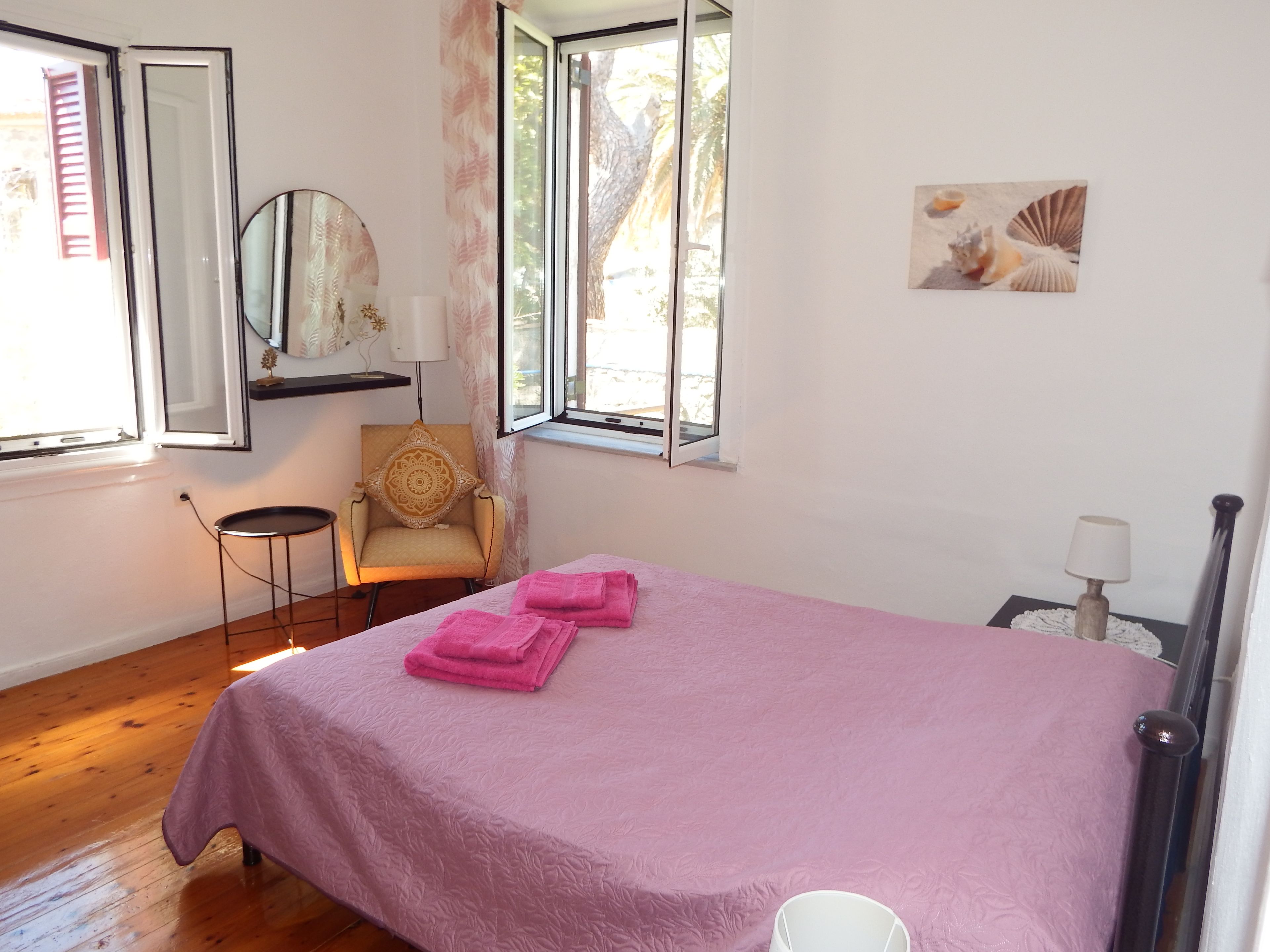 A bedroom with a bed, a chair, a window, a mirror, and a wall-mounted picture.