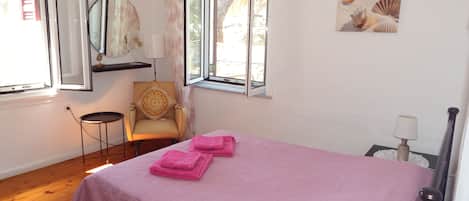 A bedroom with a bed, a chair, a window, a mirror, and a wall-mounted picture.
