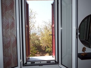 A room with a window revealing a garden view, a door with a patterned curtain, and a small plant on a shelf.