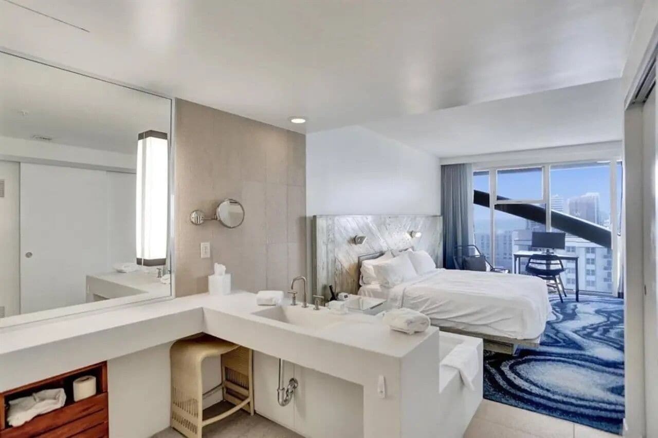 A modern hotel room with a large bed, a bathroom with a sink and mirror, and a city view through large windows.