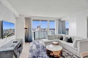 A modern living room with a large flat-screen TV, a white sofa, a round coffee table, and a city view through large windows.