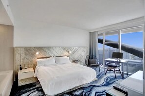 A modern hotel room with a large bed, a desk with a chair, and a view of the city through large windows.