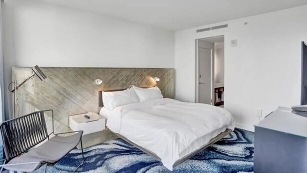 Ein modernes Hotelzimmer mit Bett, Nachttisch, Lampe, Sessel und Schreibtisch.