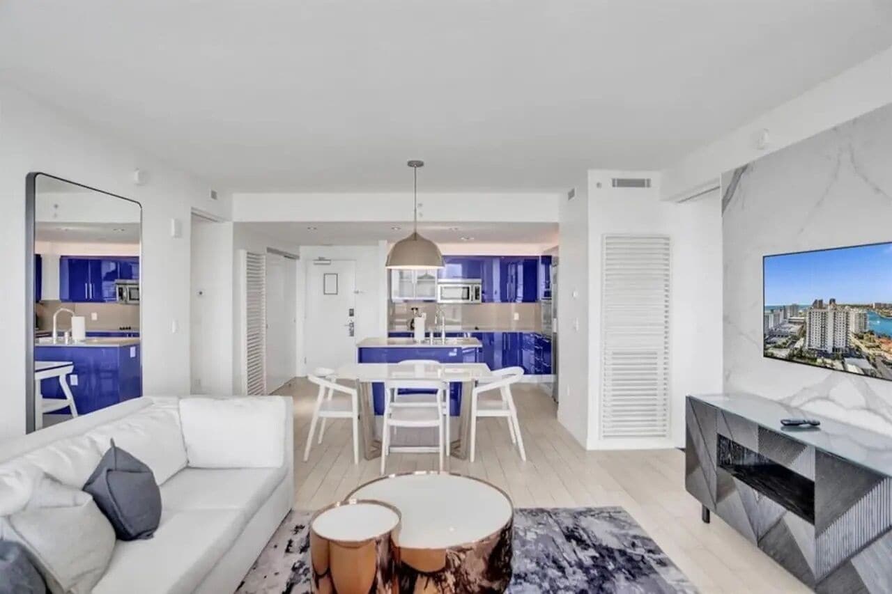 A modern living room with a sofa, coffee table, and TV. A kitchen with blue cabinets and a dining area with a table and chairs is visible.