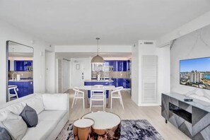 Un salon moderne avec un canapé, une table basse et un téléviseur. On peut apercevoir une cuisine avec des meubles bleus et un coin repas comprenant une table et des chaises.