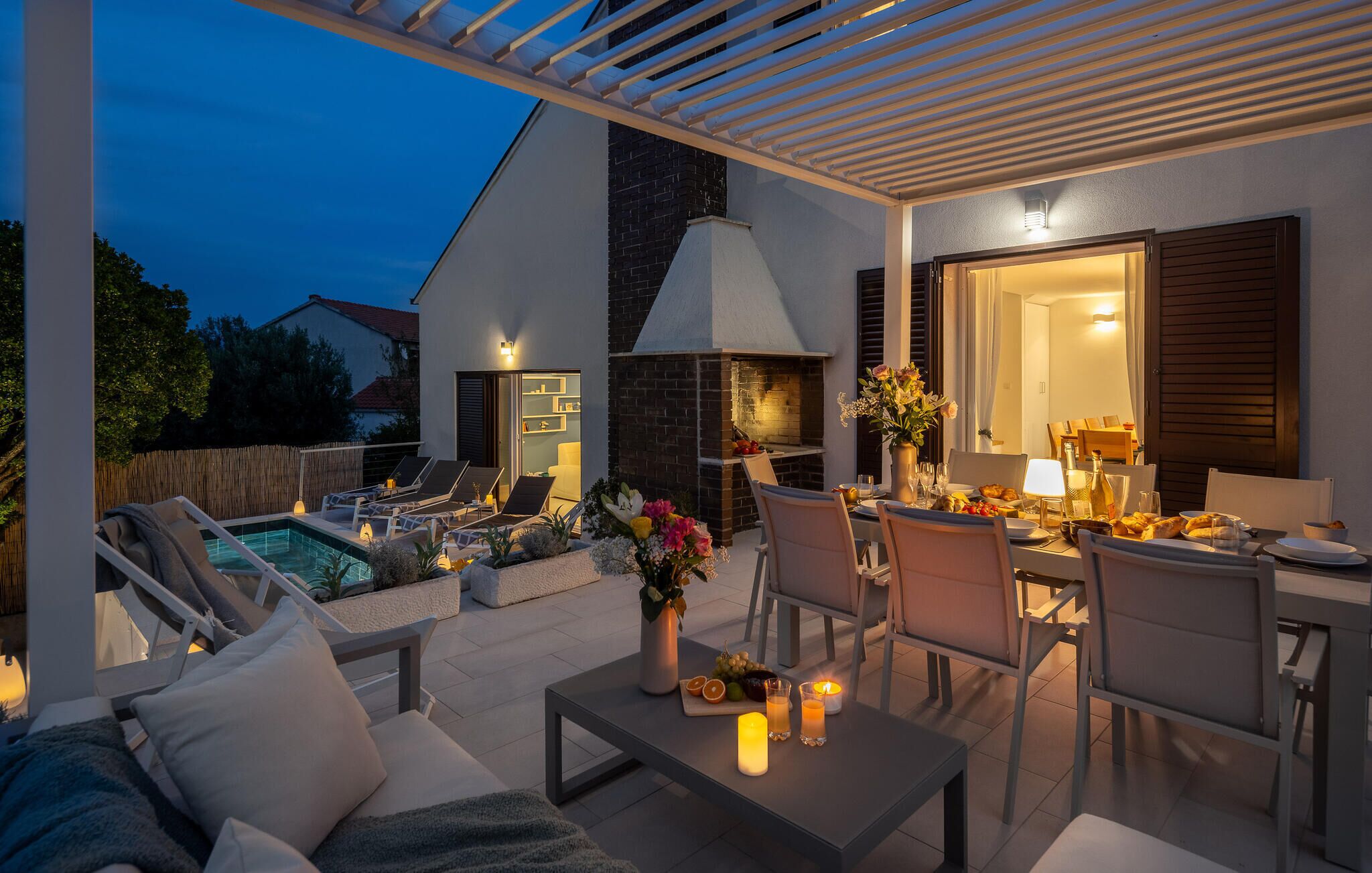 Un patio exterior bien iluminado con piscina, sillones de descanso, una mesa de comedor para dos personas y una chimenea de ladrillo.