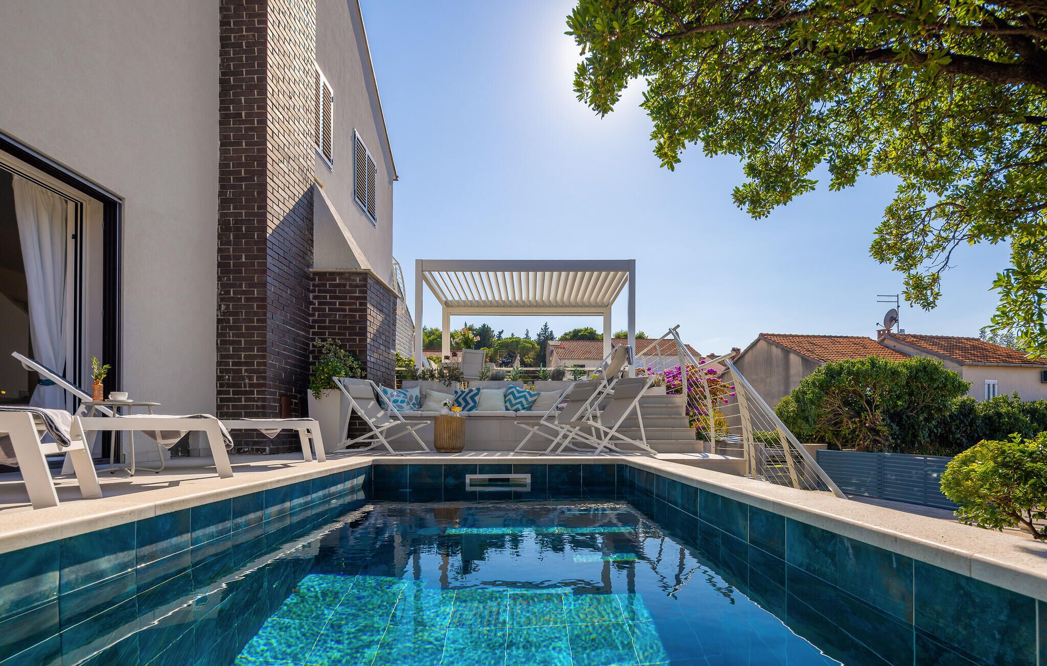A swimming pool with lounge chairs and a pergola, surrounded by greenery and buildings.