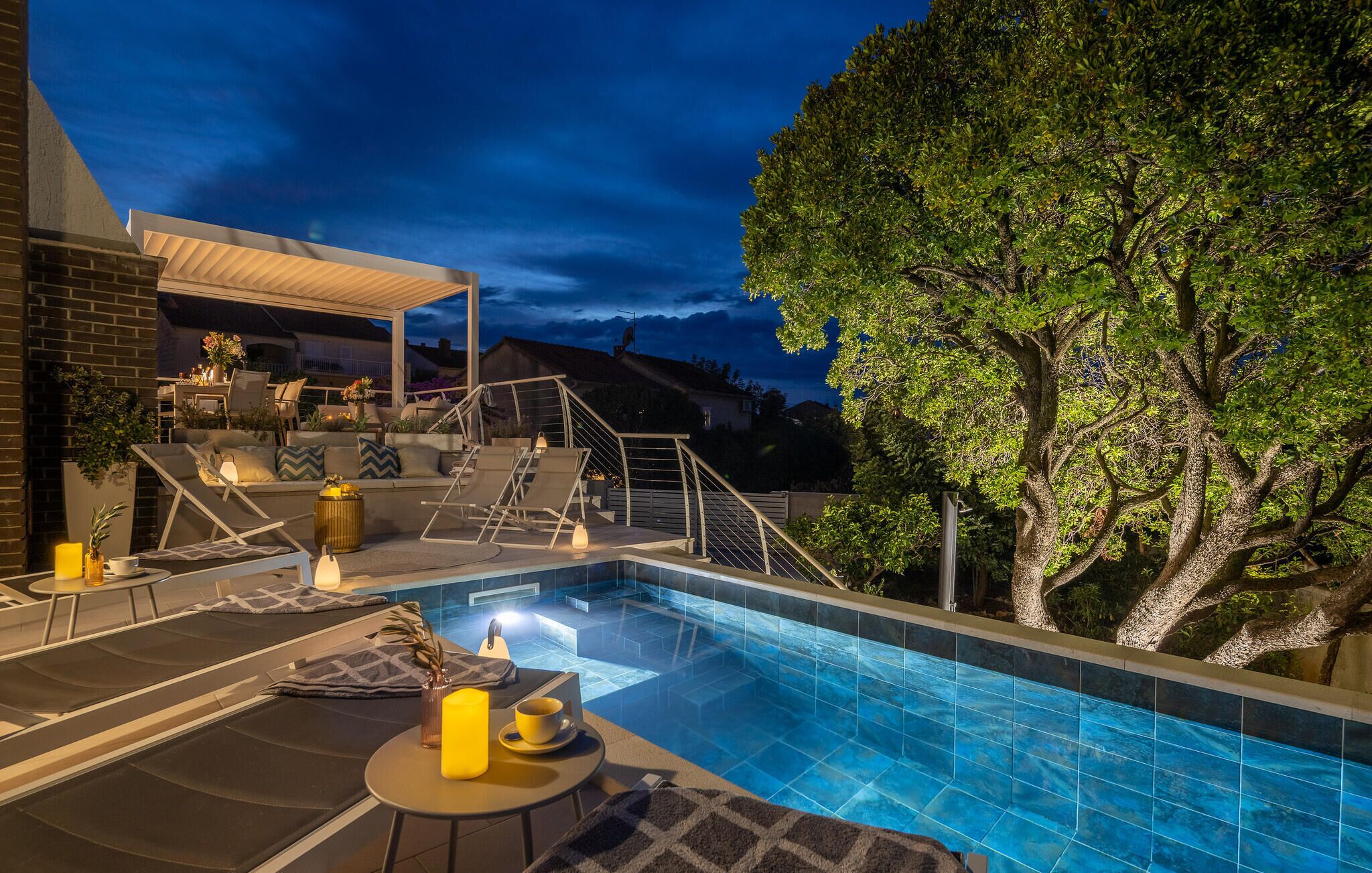 A swimming pool with lounge chairs and a covered seating area, surrounded by trees and a building.