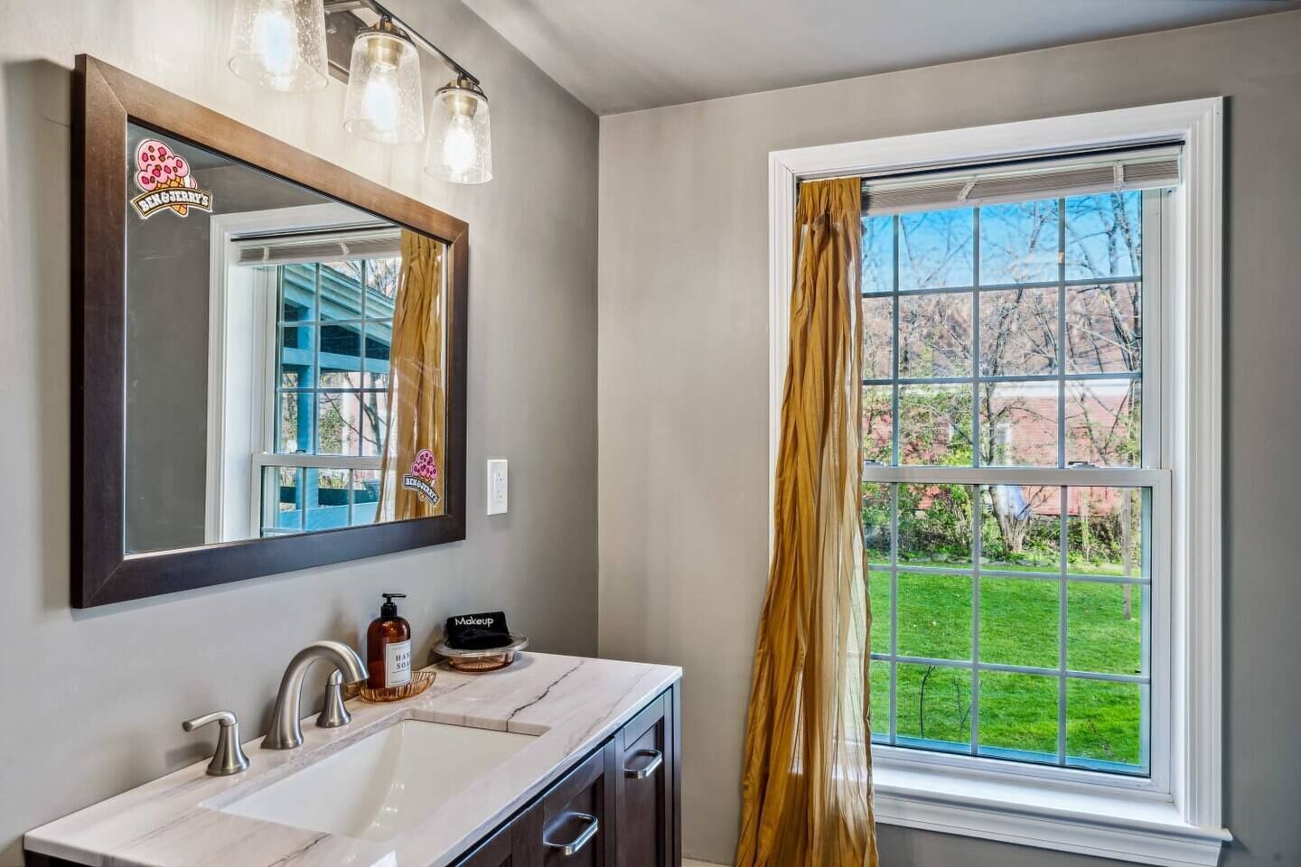 A bathroom with a large mirror, a sink, a vanity, a window with a view of greenery, and a shower curtain.