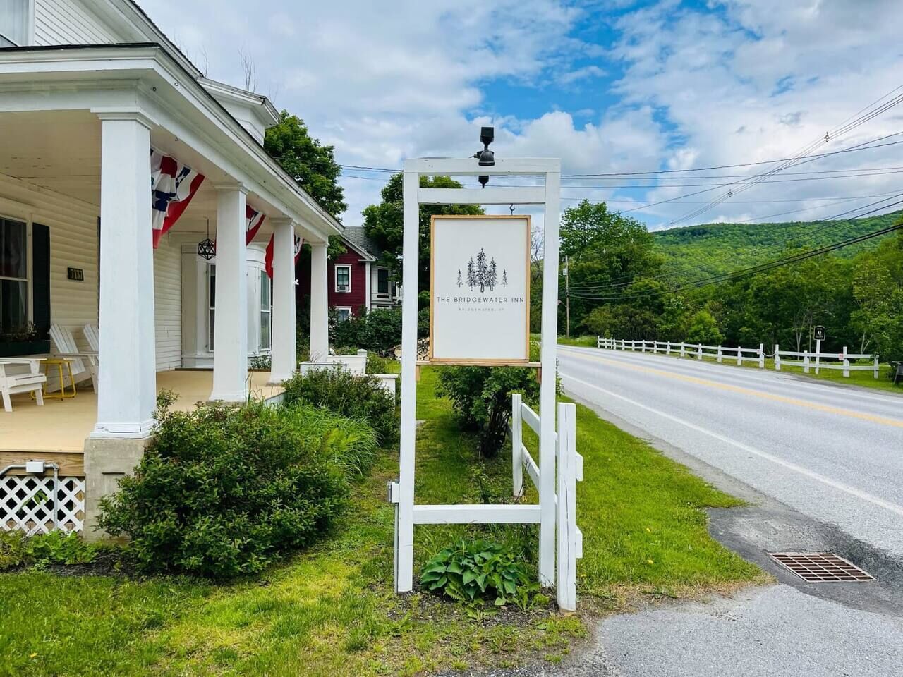 A white building with a porch, a signboard, and a road with a white fence in the background.