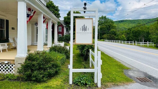A white building with a porch, a signboard, and a road with a white fence in the background.