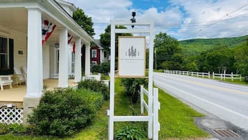 A white building with a porch, a signboard, and a road with a white fence in the background.