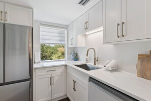 Une cuisine moderne avec des meubles blancs, un réfrigérateur en acier inoxydable, une fenêtre avec des stores et un évier avec un robinet.