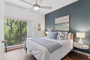 Une chambre avec un grand lit, un ventilateur de plafond, une vue sur les arbres par la fenêtre et un tableau au mur.