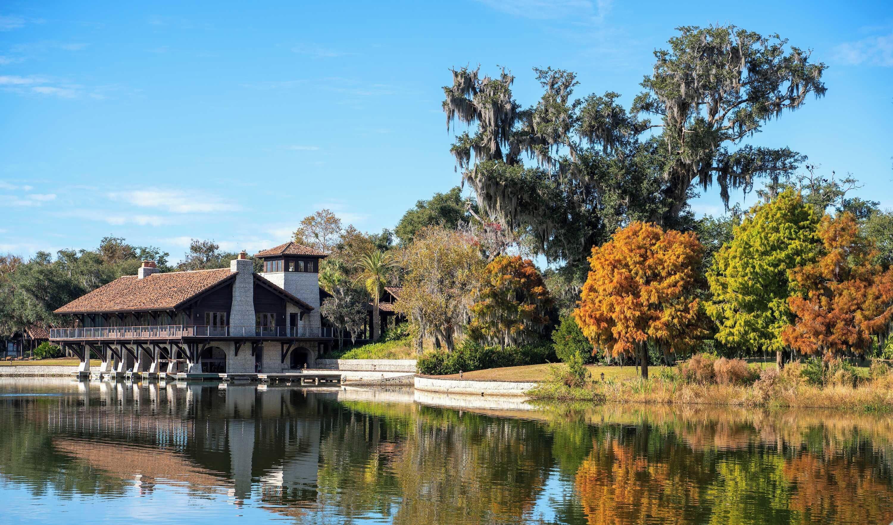 Un edificio histórico con fachada de piedra y techo de tejas, ubicado junto a un lago tranquilo que refleja árboles y el edificio.