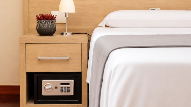 A neatly made bed with a white comforter and a grey blanket, a bedside lamp, a small decorative plant, and a nightstand with a drawer and a built-in safe.