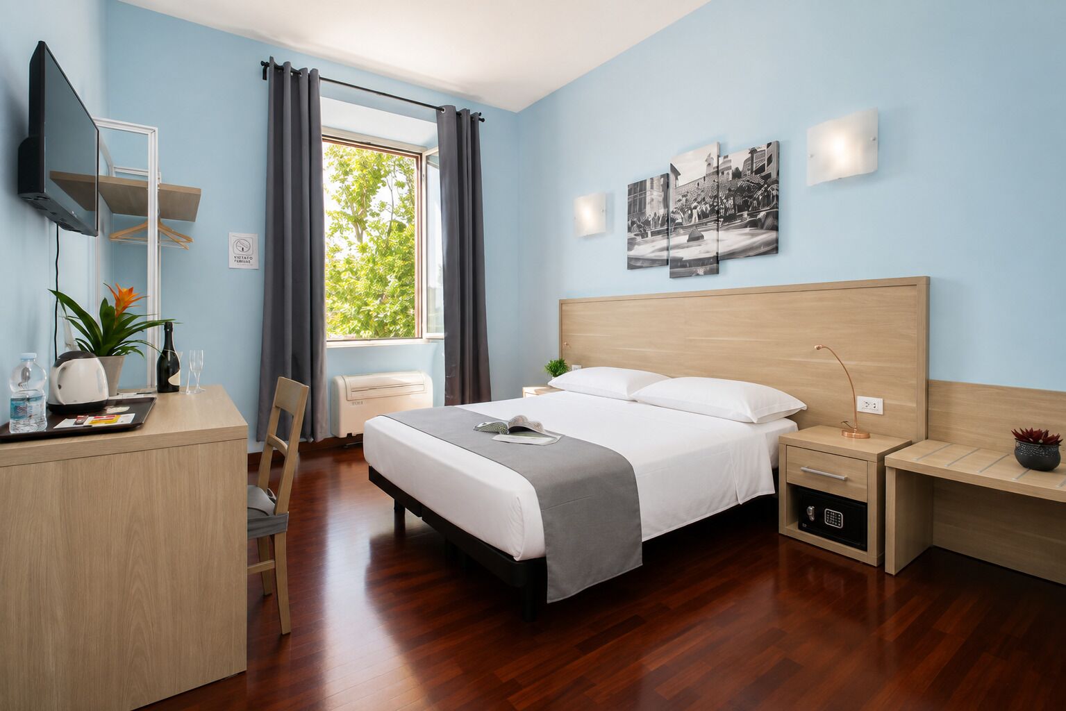 A hotel room with a bed, bedside tables, a desk, a chair, a TV, a window with curtains, and a wall-mounted picture.