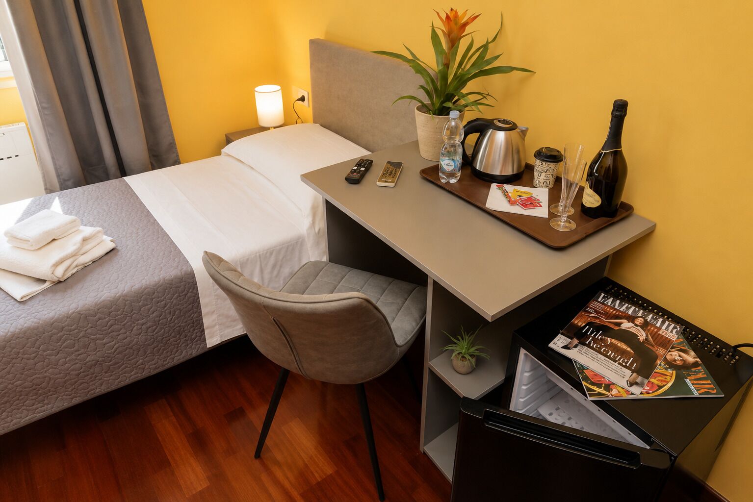 A hotel room with a bed, a desk with a chair, a small table with a plant, a kettle, a bottle, and a magazine.