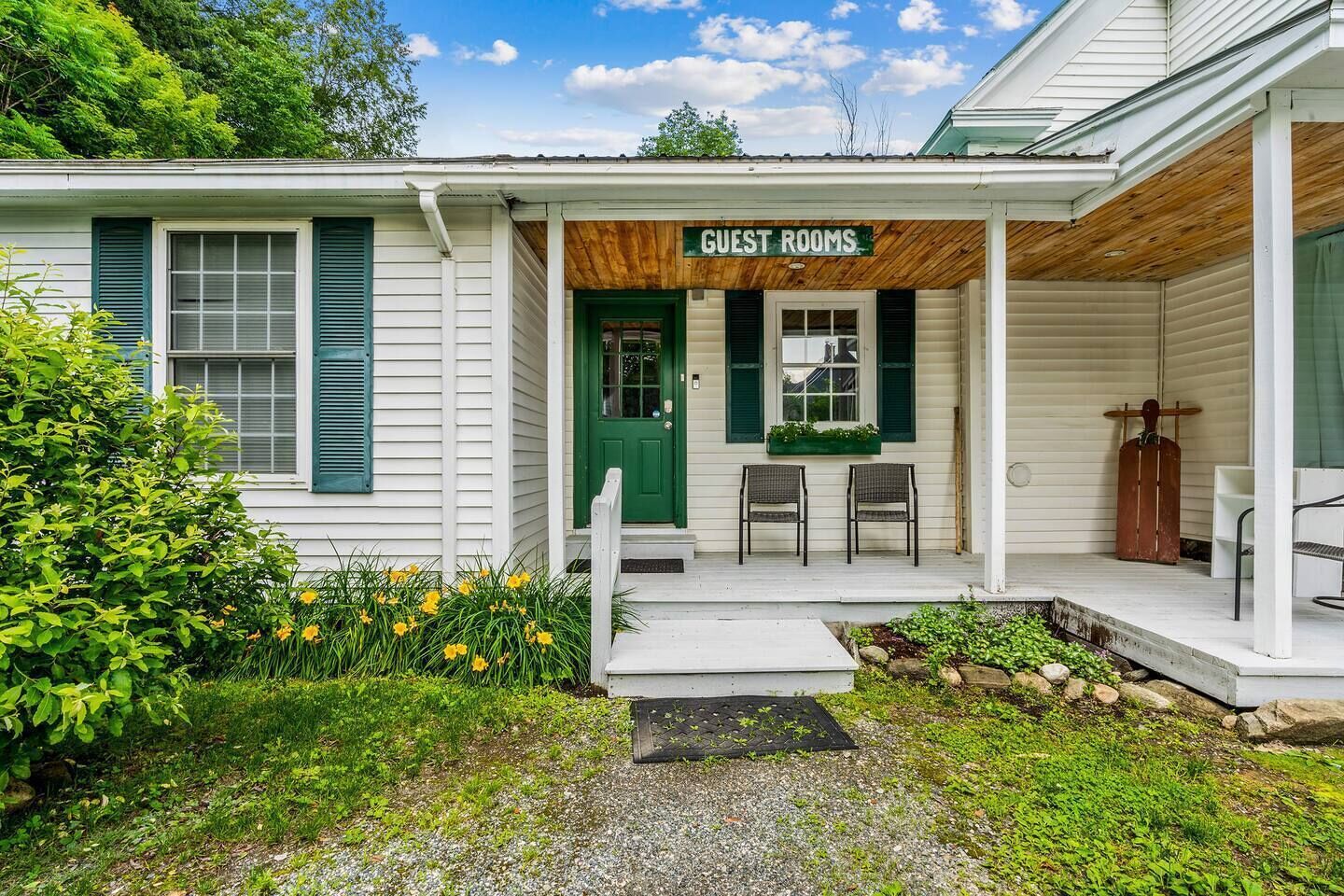 A small building with a porch labeled 'GUEST ROOMS,' two chairs, a green door, and a wooden bench.