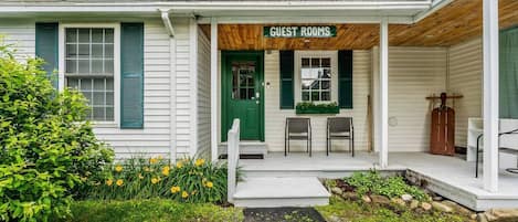 Un pequeño edificio con un porche que indica 'GUEST ROOMS', dos sillas, una puerta verde y un banco de madera.
