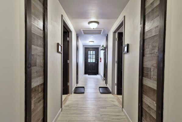 Un pasillo de hotel con suelo de madera clara, paneles de madera oscura en las paredes, una puerta negra al final y un extintor montado en la pared.