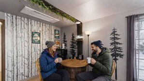 Two people seated at a round wooden table in a room with a forest-themed wallpaper, a coffee machine, and a window with curtains.