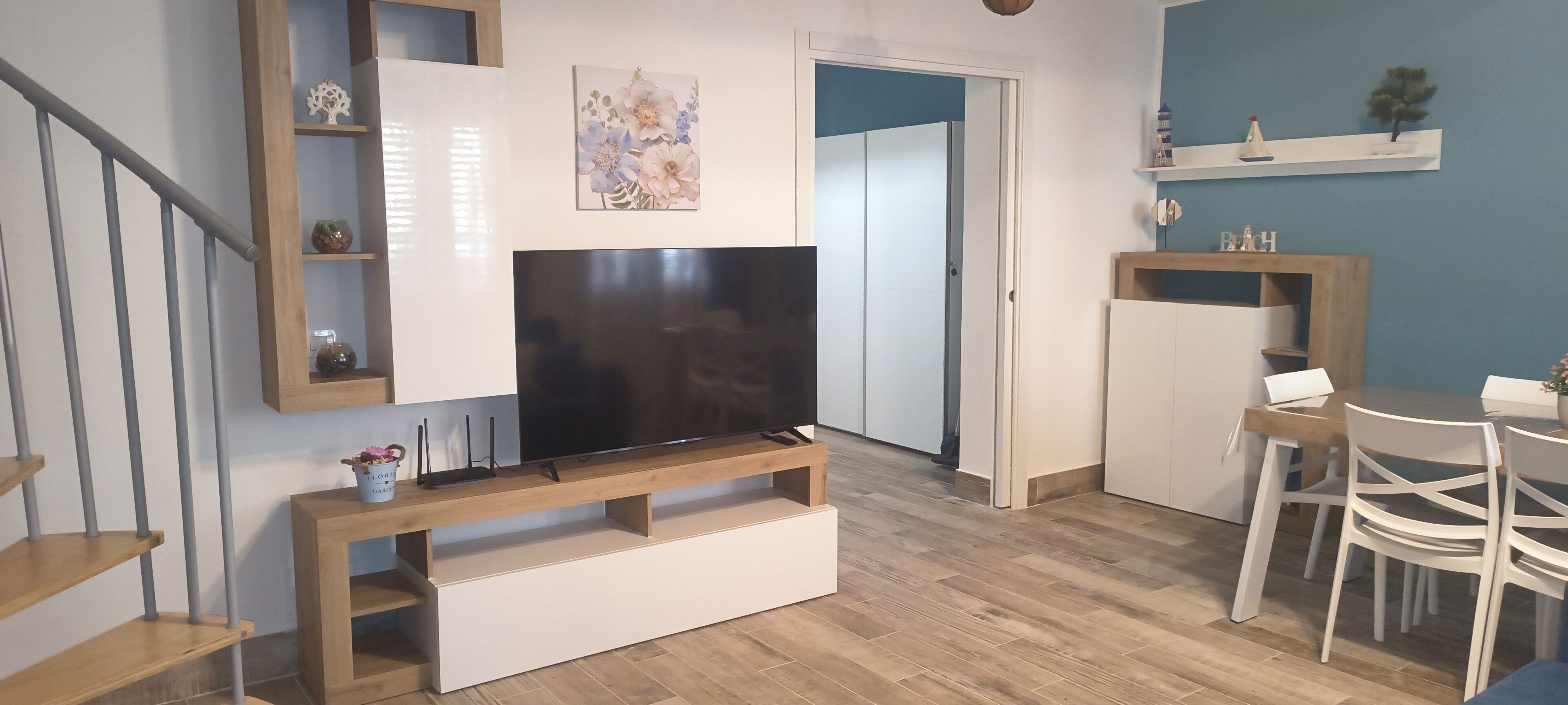 Uma sala de estar com uma TV de tela plana em um suporte de madeira, uma área de jantar com mesa e cadeiras, e uma escada com corrimão.