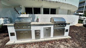 Ein Grillbereich im Freien mit zwei Gasgrills und einem integrierten Kühlraum, gelegen in einer begrünten Anlage mit Mulchschicht.