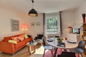 Een moderne woonkamer met een bank, een eettafel, stoelen, een televisie en een raam met gordijnen.