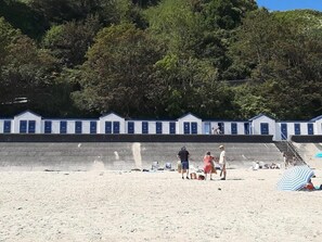 Una playa con una hilera de edificios blancos con puertas azules, una zona arenosa con gente y una colina boscosa al fondo.