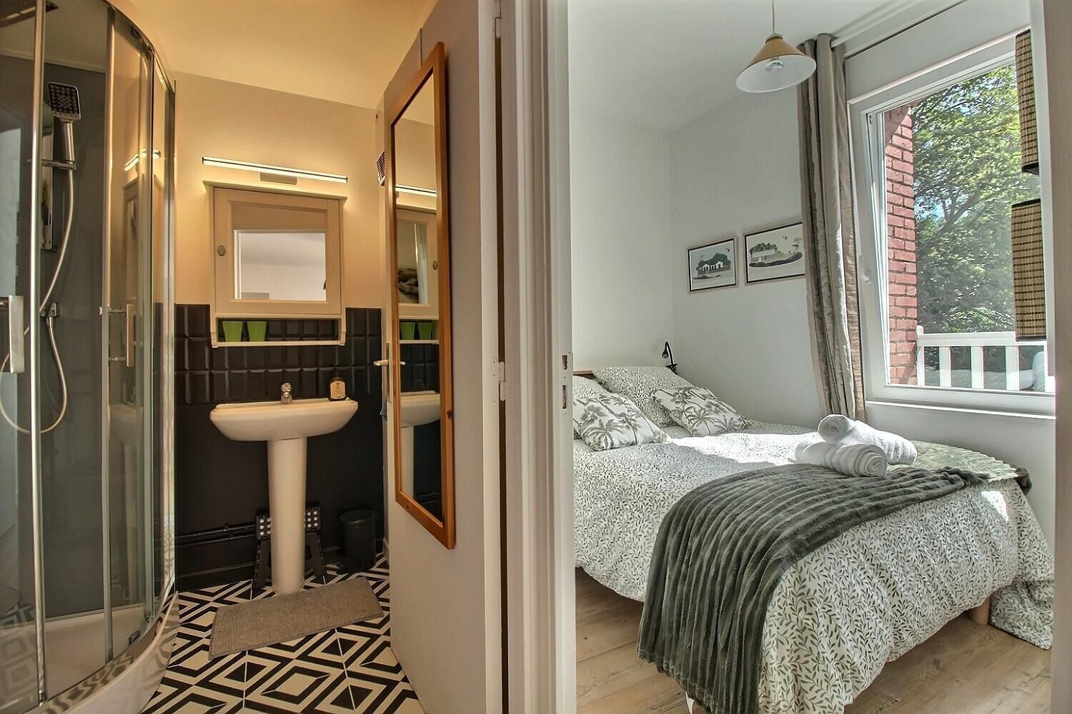 A bathroom with a shower, sink, and mirror, and a bedroom with a bed, window, and artwork.